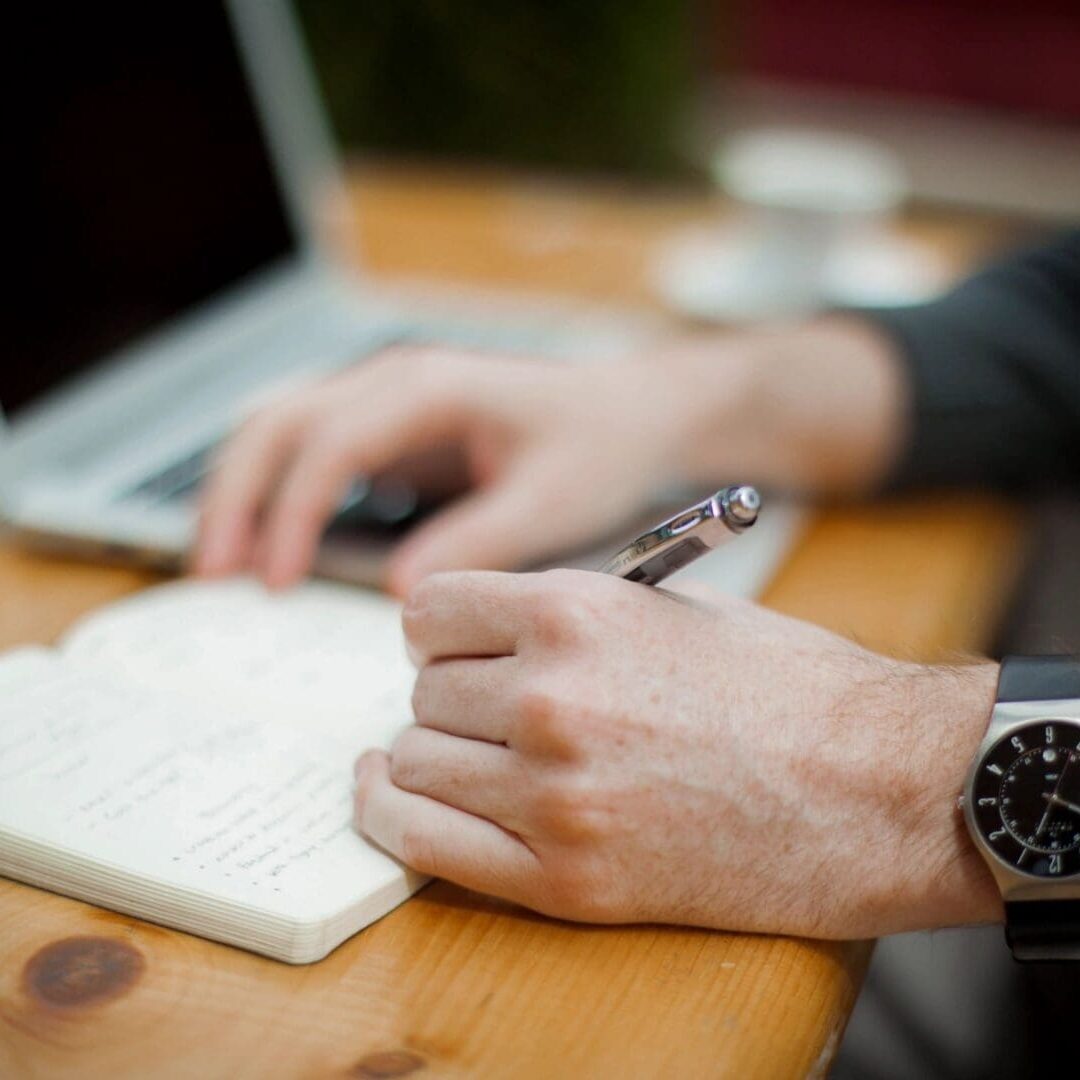Person writing in notebook near laptop.